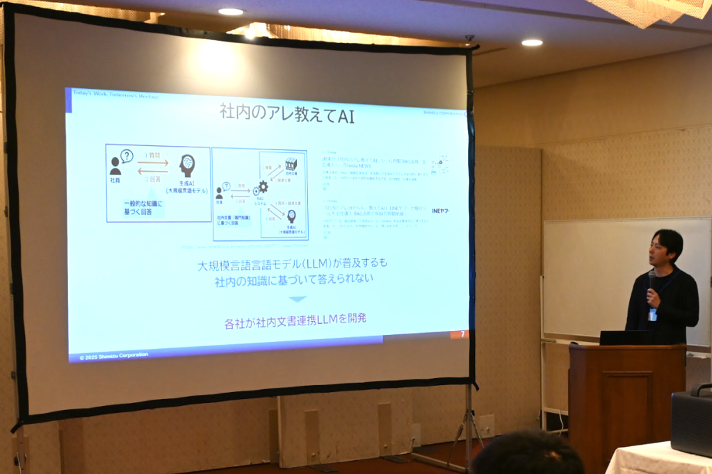 清水建設による発表スライドで、「社内のアレ教えてAI」というタイトルの下に、社内知識を活用したAIシステムの概念図が表示されている。スライドには「大規模言語言語モデル(LLM)が普及するも社内の知識に基づいて答えられない」という課題と、「各社が社内文書連携LLMを開発」という解決策が示されている。講演者が壇上で説明している様子。