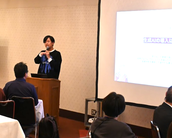 壇上で黒いシャツに青いロゴが入った衣装を着たおざけん氏がマイクを持ち、生成AI活用の事例について講演している様子。木製の演台に立ち、聴衆に向かって手振りを交えながら説明している。前列には参加者が着席して聞き入っている。