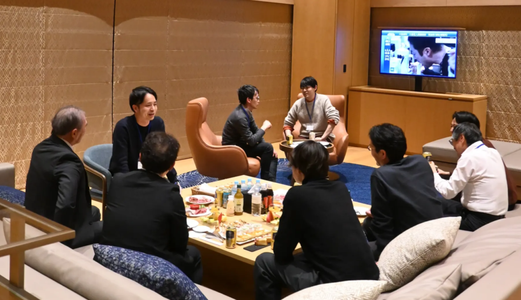 安比リゾート内のラウンジでの懇親会の様子。参加者が円形に配置されたソファとテーブルを囲み、飲食しながら交流している。テーブル上には様々な飲み物や軽食が並び、参加者はリラックスした雰囲気の中で会話を楽しんでいる。奥にはテレビモニターが設置されている。ビジネスカジュアルな装いの参加者たちが和やかに意見交換している。